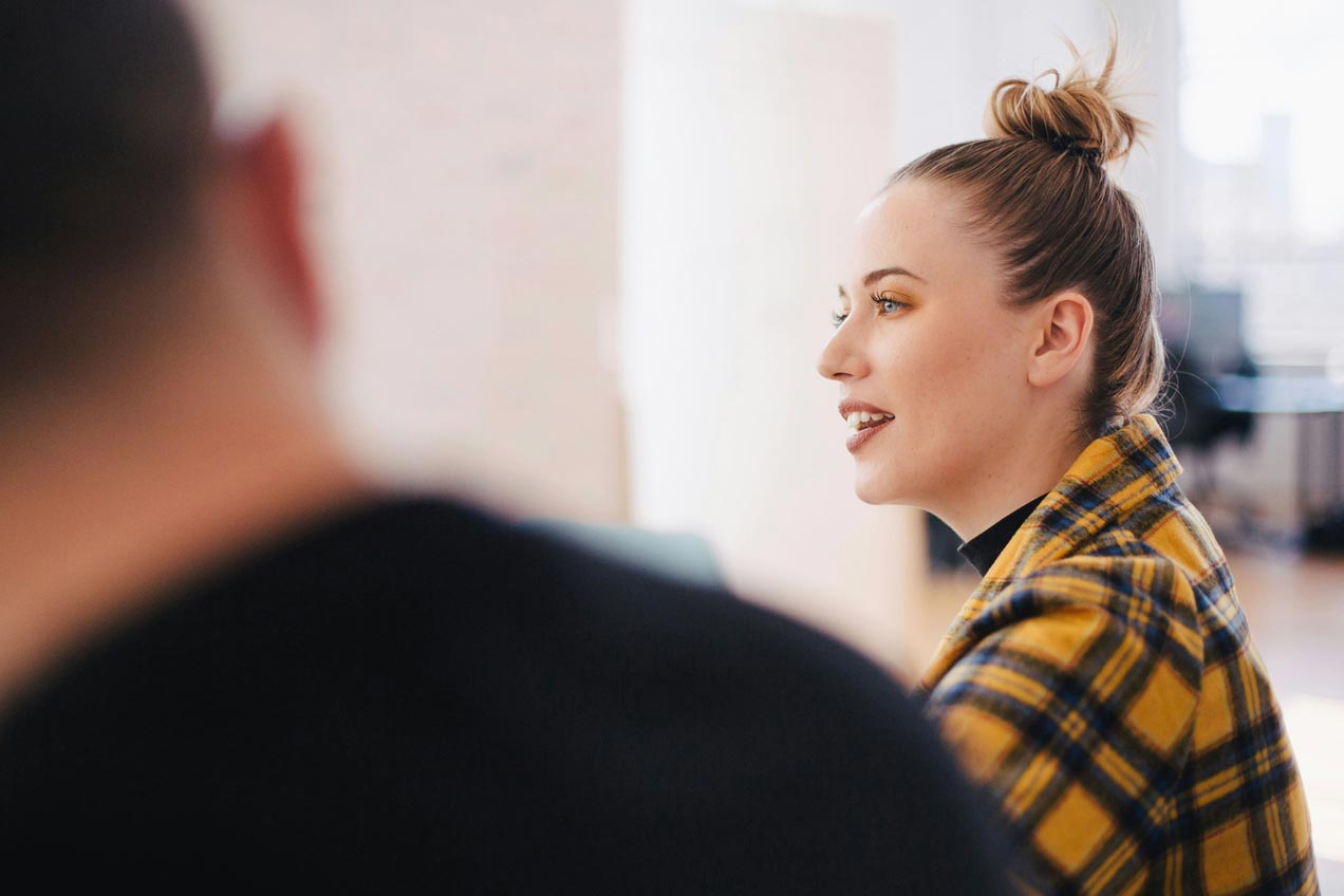 A woman wearing a yellow plaid shirt and a topknot hairstyle is speaking in a well-lit room. A blurred person in the foreground listens.