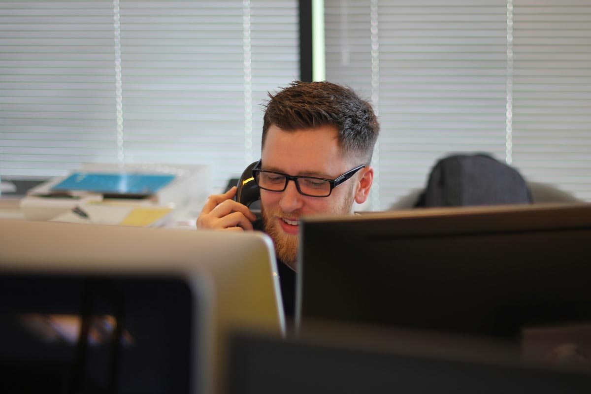 A man wearing glasses sits behind two computer monitors. He is holding an office phone to his ear while looking down a something in front of him.