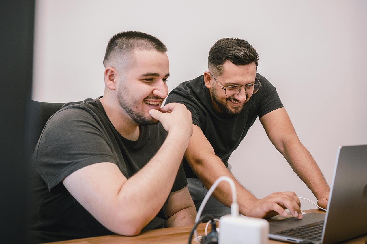 Two men are smiling and interacting with a laptop at a desk, with one sitting and the other standing.