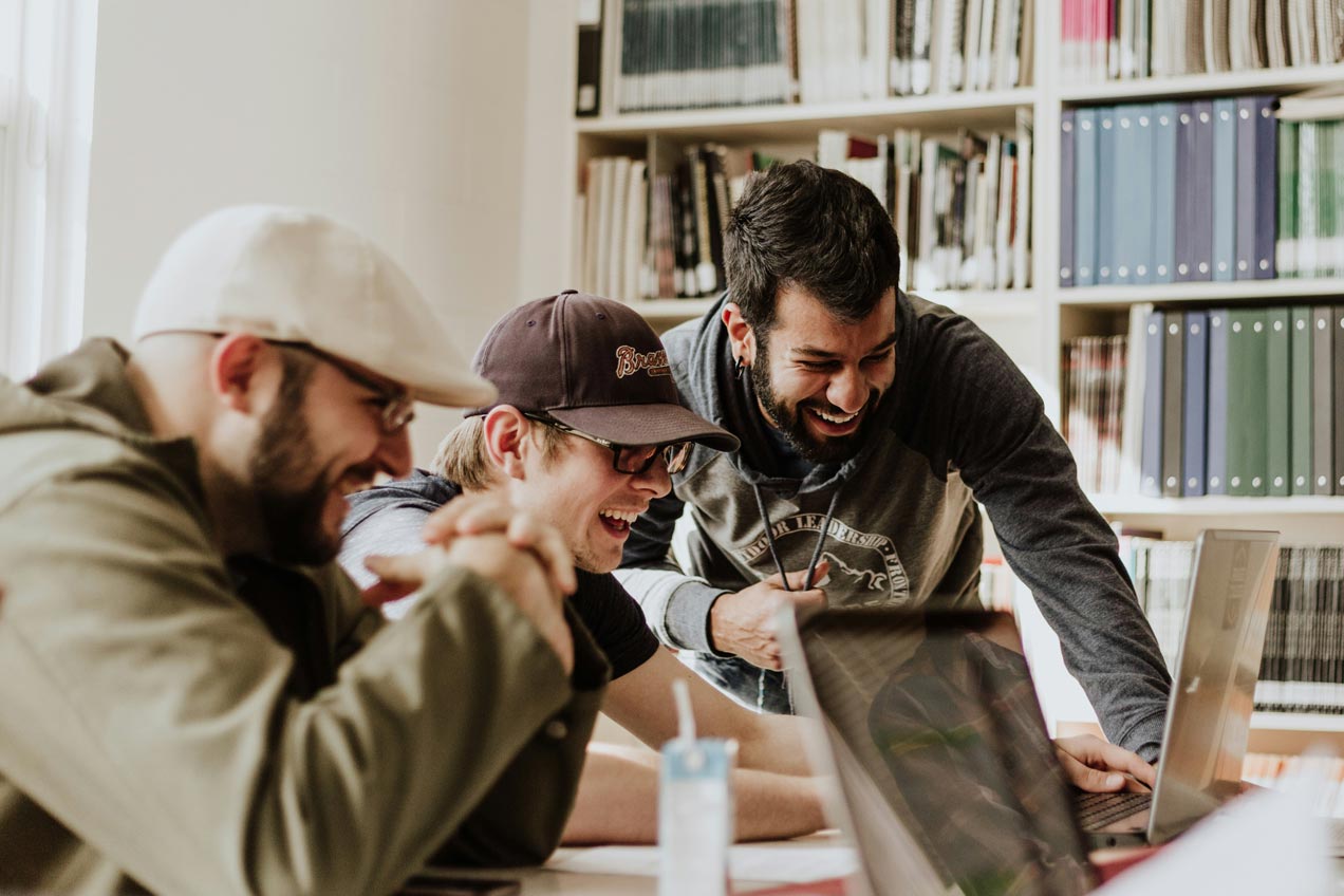 Three men sitting at a table, smiling and looking at a laptop screen. Bookshelves are in the background.