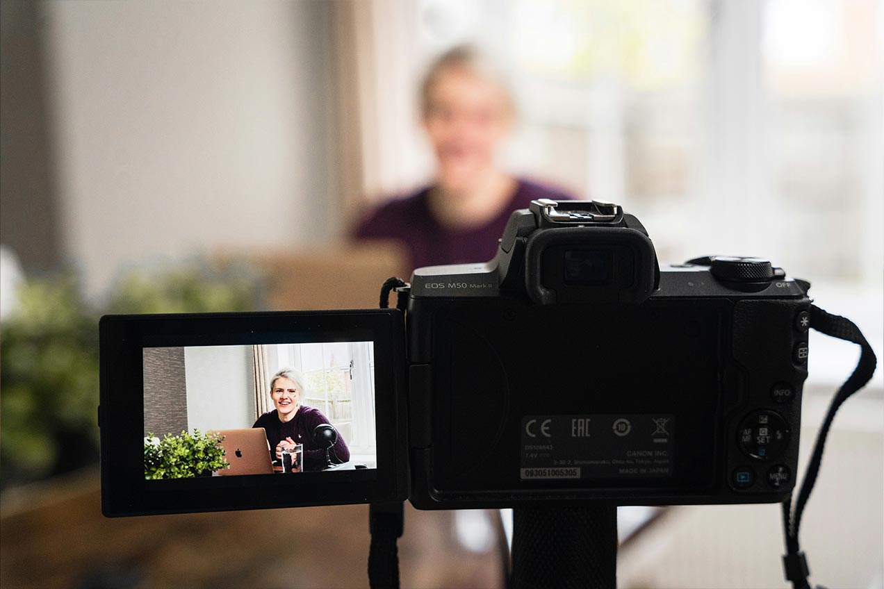 A camera is focused on a person appearing on the screen. The person is smiling and sitting indoors in front of a blurred background.