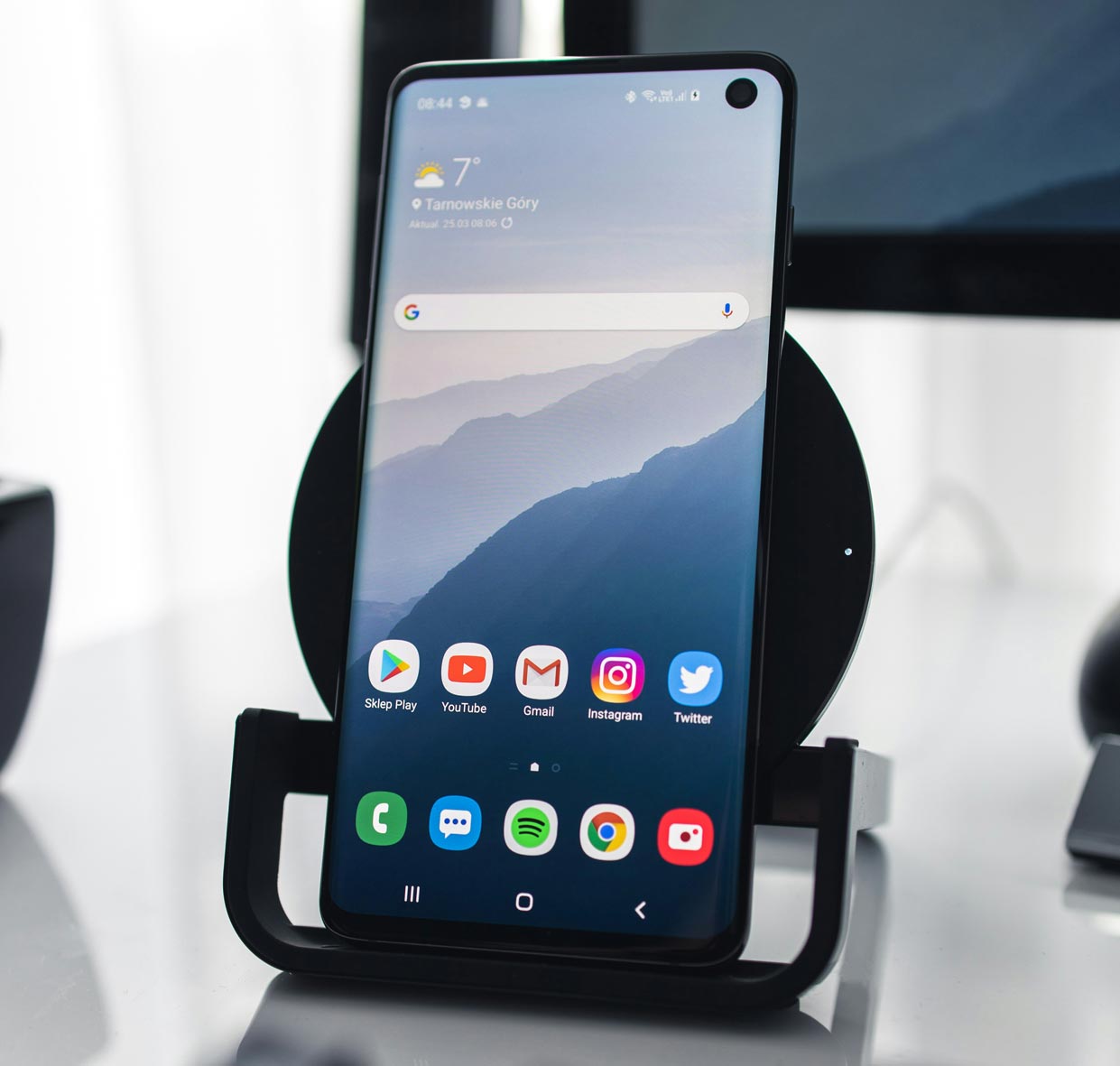 A close up shot of an Android smartphone sitting in a phone holder on a white desk. The phone screen shows the home screen and has blue, misty mountains as the background wallpaper.