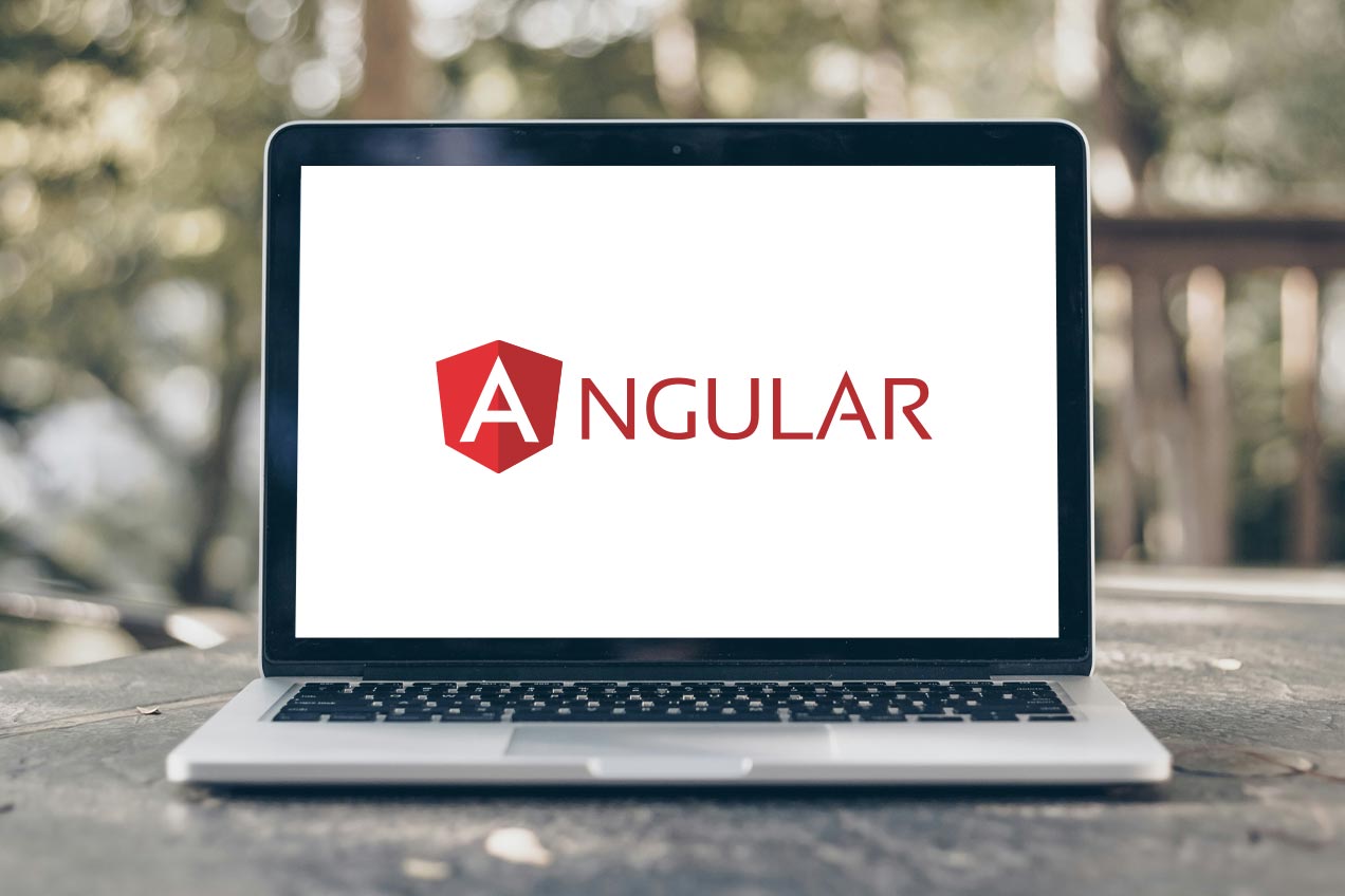 A laptop on a wooden table outdoors displays the Angular JS logo on its screen, with blurred greenery in the background.