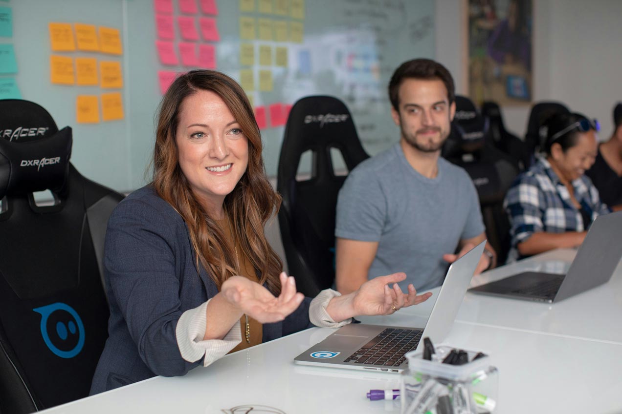 A row of software developers sitting at a long white desk, each with open laptop in front of them. The person nearest the camera is gesturing with her hands to someone off camera.