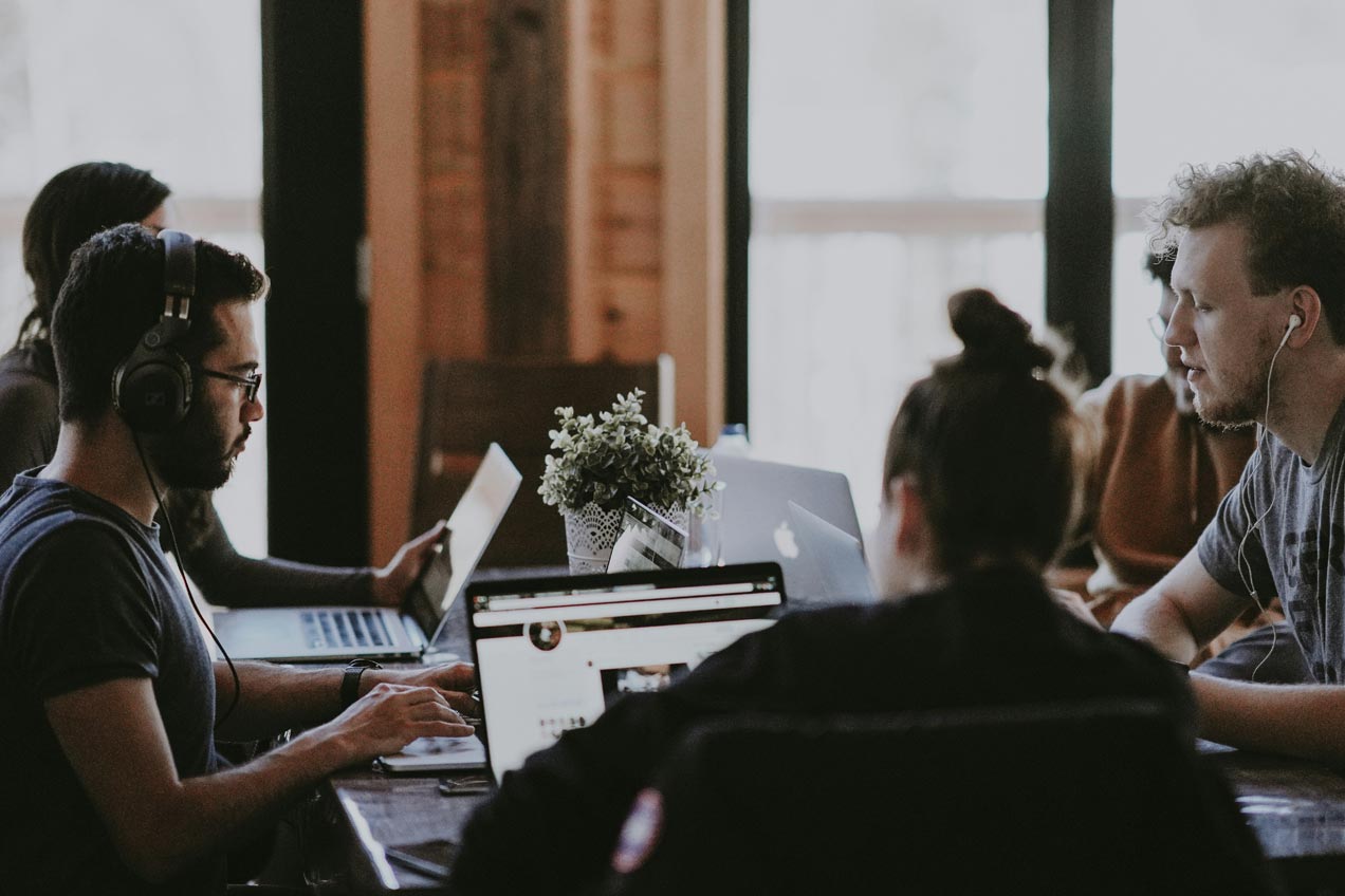 A group of five software developers are sitting round a wooden table, each of them working on laptopns. They all are concentrating on their work, with two of them wearing headphones and earphones.