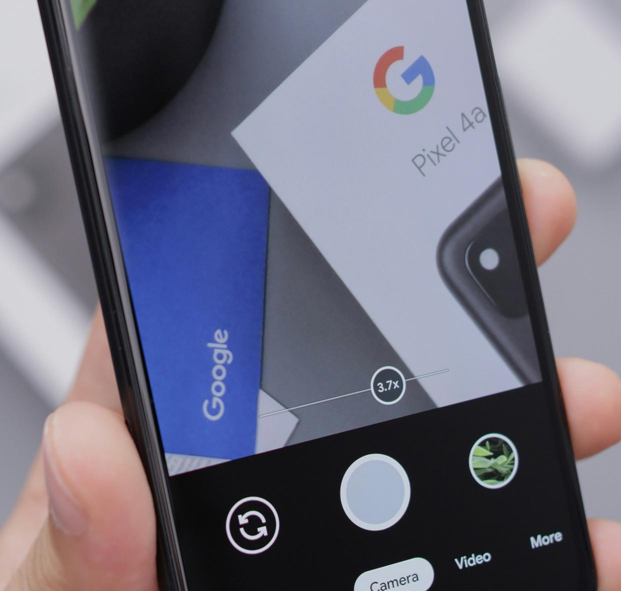 A close up of a Google Pixel smartphone being held in someone's hand. The screen shows the camera app open and focusing on Google Pixel 4a packaging.