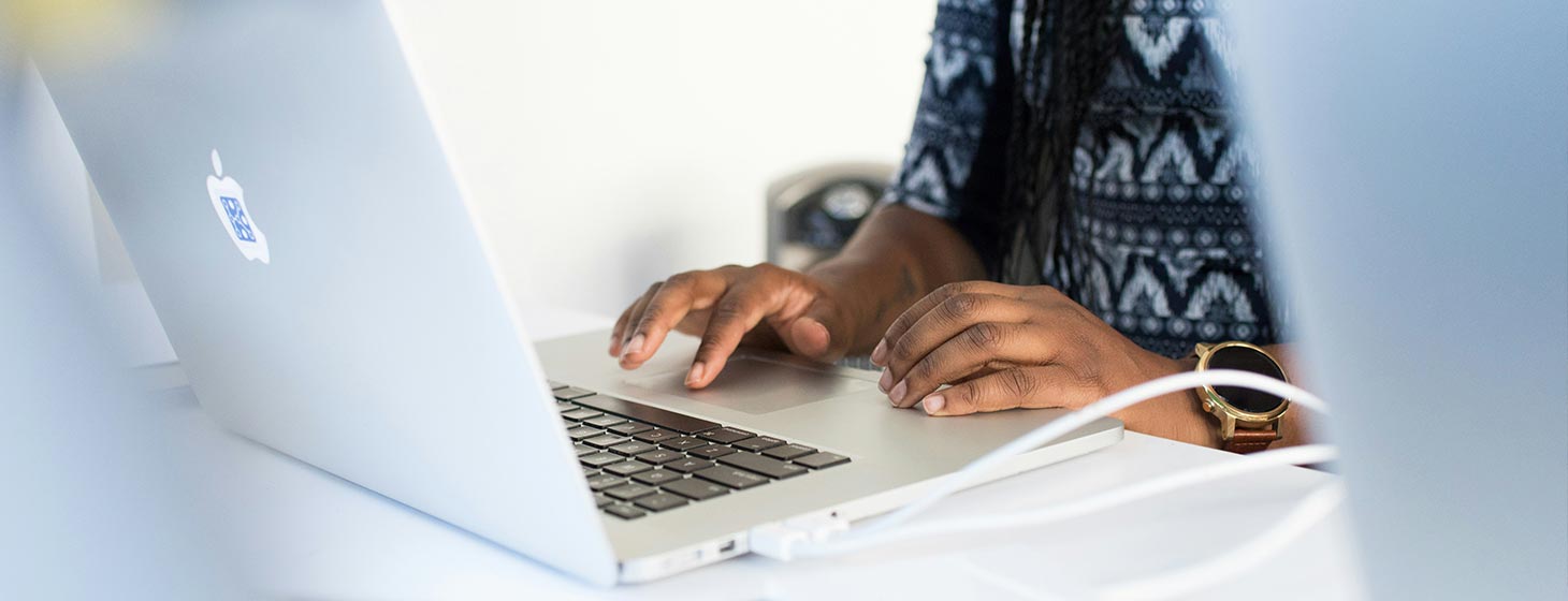 A close of a person sitting working at a white desk. Their hands are resting on an open laptop.