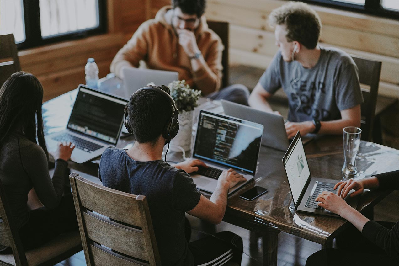 A group of five software developers are sitting round a wooden table, each of them working on laptops. They all are concentrating on their work, with two of them wearing headphones and earphones.