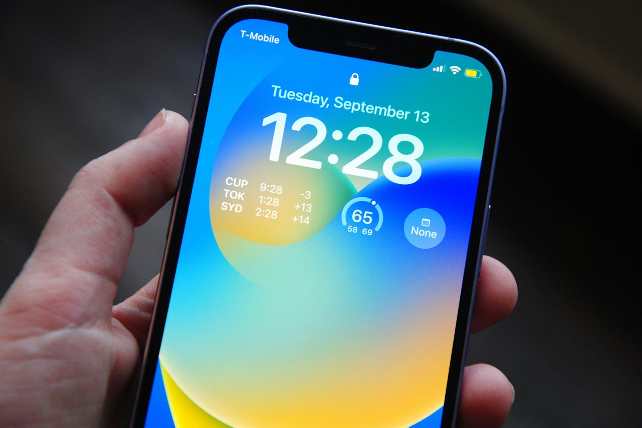 A close up shot of an iPhone being held in someone's hand. The phone screen shows the home screen and has a blue, yellow and green abstract background wallpaper.