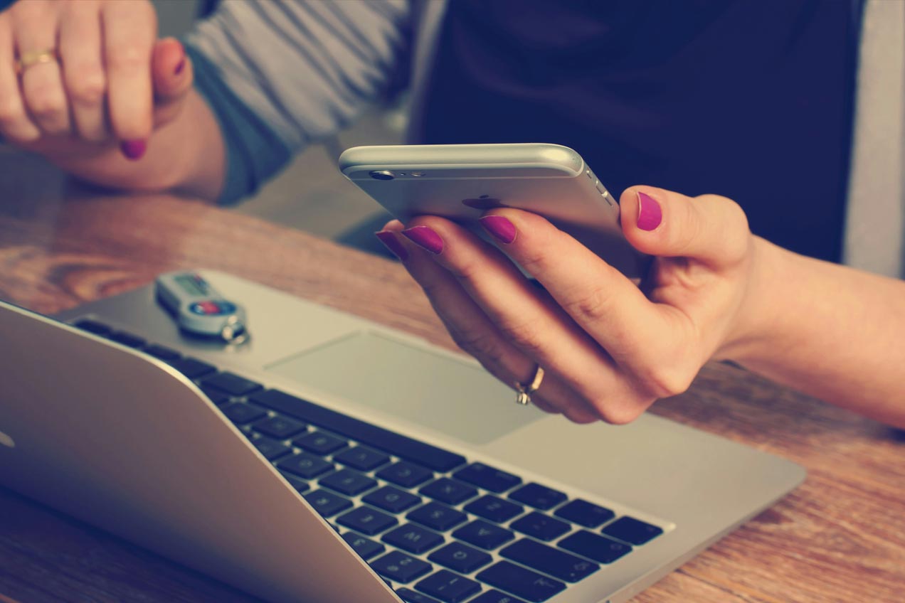 A close up shot showing a person sitting at a wooden desk with a laptop open. In their left hand they are holding an iPhone. To the right of the trackpad on the laptop sits a USB storage device.