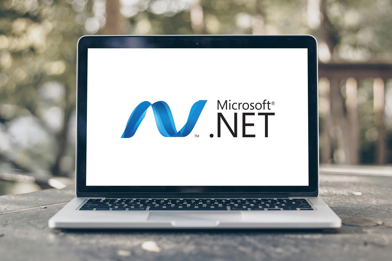 A laptop on a wooden table outdoors displays the Microsoft .NET logo on its screen, with blurred greenery in the background.