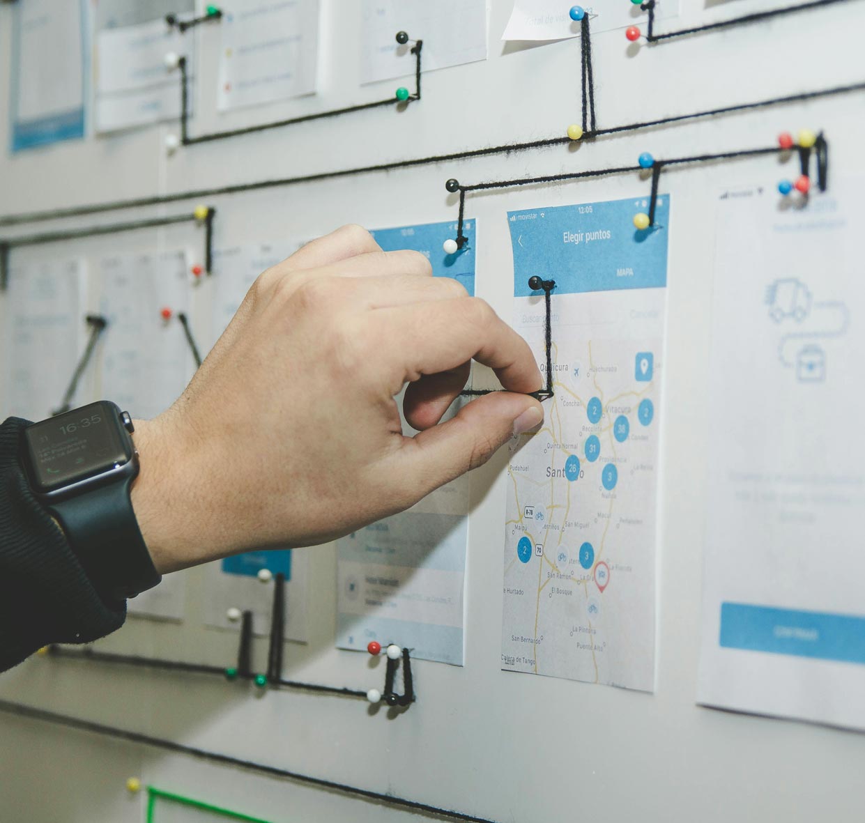 Close up of a person's hand connecting string between pins on a wall of mobile app designs.