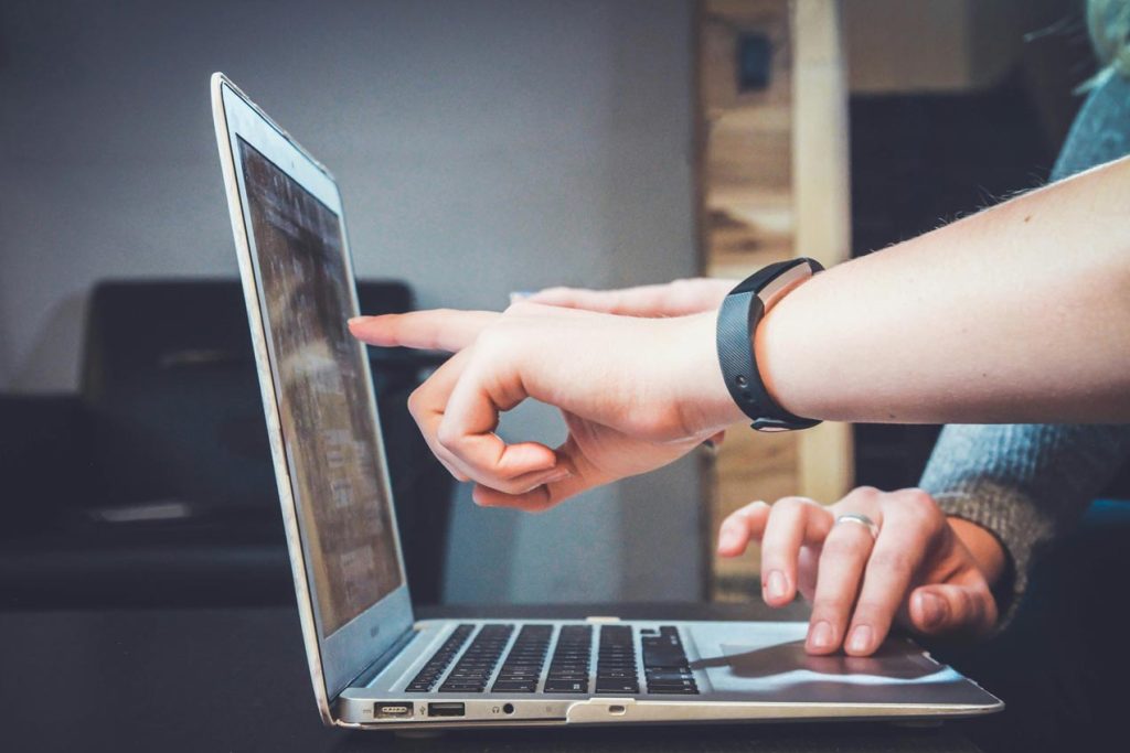 A person points at a laptop screen while sitting at a desk, wearing a fitness band and a ring, suggesting a work or study environment.