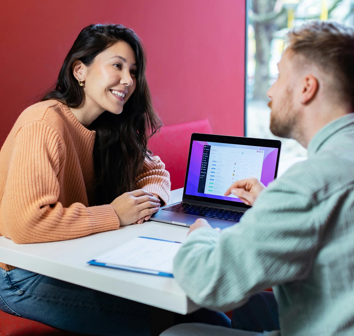 Two people are having a conversation at a table with a laptop showing a spreadsheet. They're in a casual, colorful setting, possibly collaborating or studying.