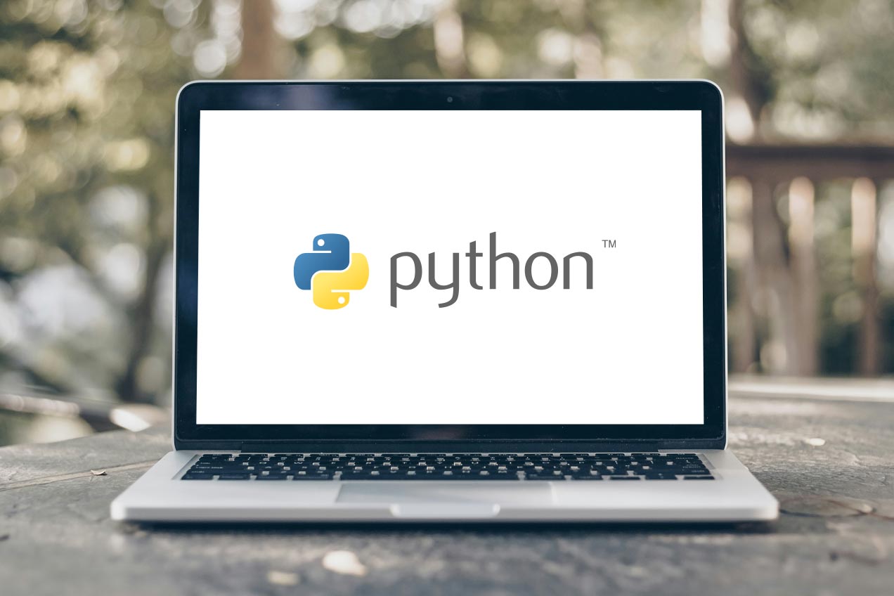 A laptop on a wooden table outdoors displays the Python logo on its screen, with blurred greenery in the background.