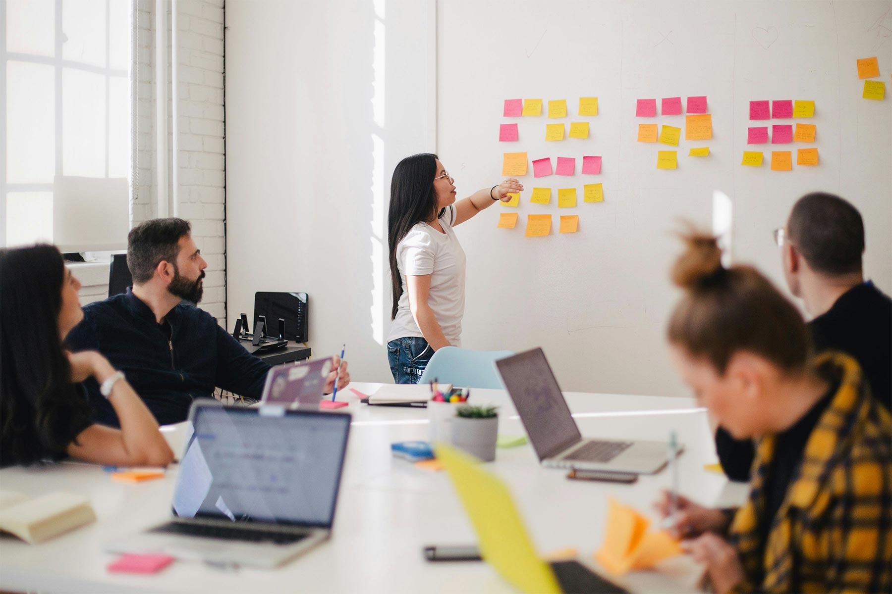 Four software developers are sitting round a large white table, each of them with laptops open in front of them. A fifth person is standing looking at a wall that has lots of brightly coloured post it notes stuck to them.