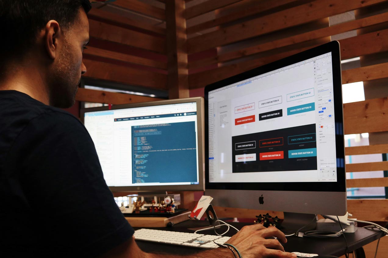 A web developer is sitting at a desk in front of two computer montitors. The left monitors shows CSS code, while the right monitor show different colour variations ans states of user interface button designs.