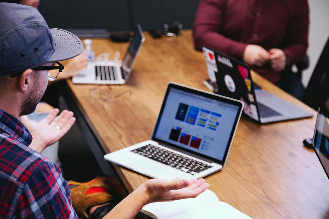 Three people in a casual office setting with laptops discussing graphs displayed on a screen, likely collaborating or reviewing work. One person gestures.